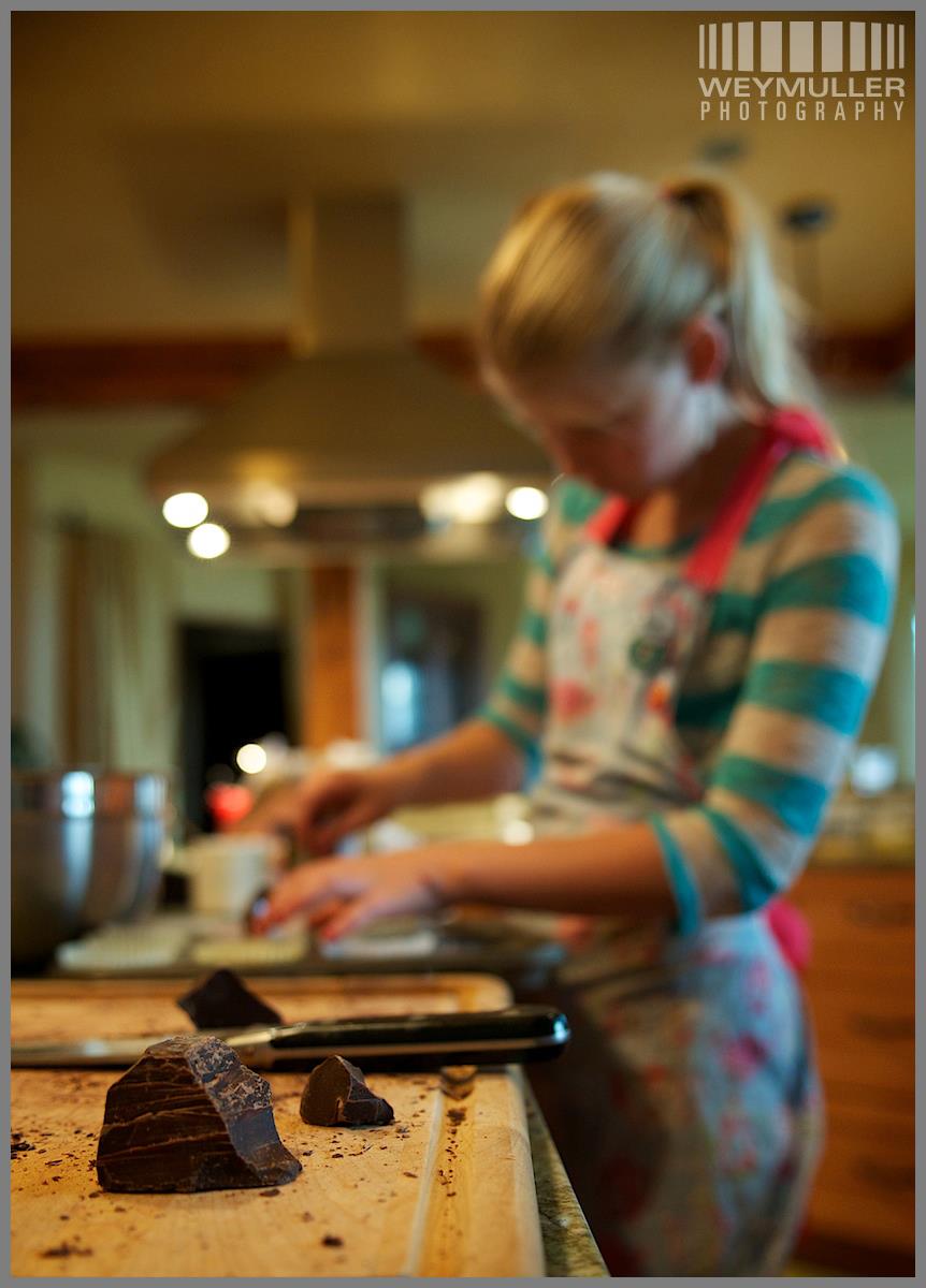 Young Hannah in a teal striped shirt and floral apron, breaking chocolate on a wooden cutting board. Photo by Weymuller Photography.