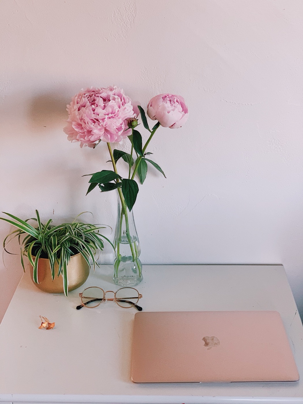 A workspace with pink peonies in a glass vase, a rose-gold laptop, round glasses, and a small spider plant in a gold pot